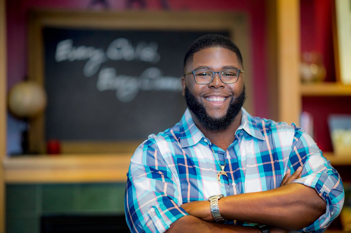 Anthony Abraham Jack, an african-american man with a full beard and glasses, smiles, standing with crossed arms in front of a blackboard.