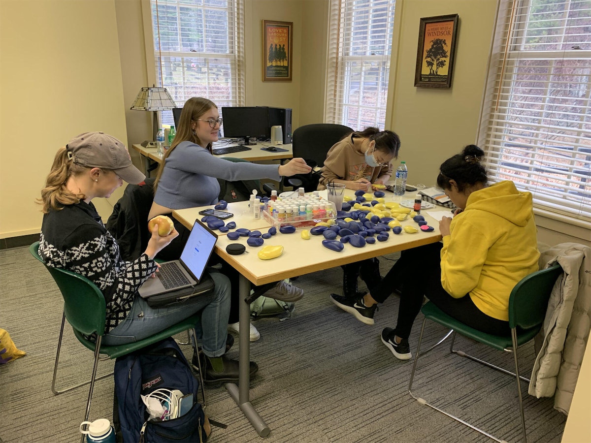 Students gathered around a table paint rocks in maize and blue colors.
