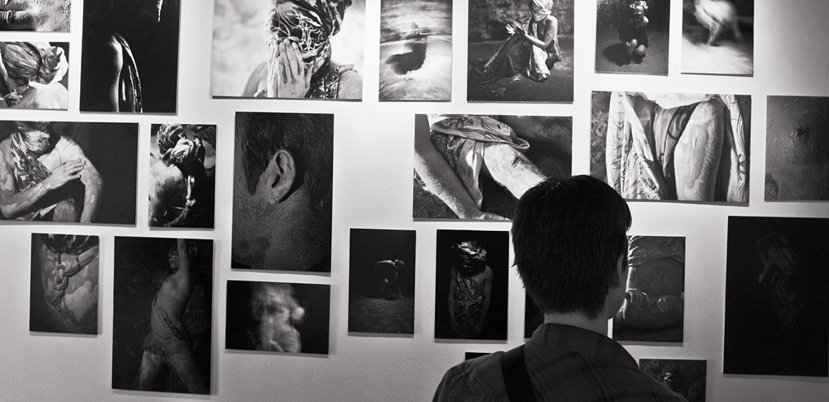 A figure, shown from behind, examines photographs hanging on a wall in this black and white image